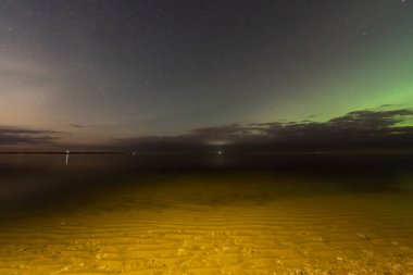 Estonya sahillerinde sakin bir gece manzarası, yıldızlı gökyüzü, yeşil aurora borealis, ve sakin Baltık Denizi suları.