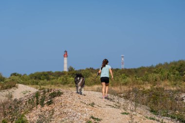 Genç kız ve Sibirya Husky köpeği deniz feneri geçmişi olan Estonya 'da manzaralı bir sahil yolunda yürüyüş yapıyorlar..