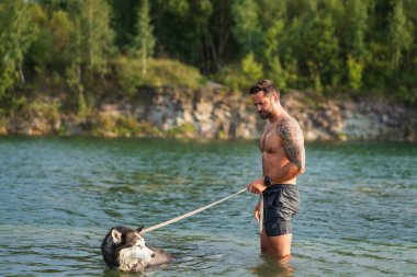 Kaslı, dövmeli bir adam güneşli bir günde sadık Sibiryalı Husky köpeğiyle yenilenen bir yüzüşten zevk alır..