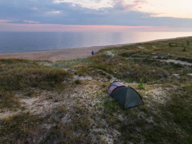Estonya kıyı kum tepelerindeki çadırlarından, Baltık Denizi üzerindeki sakin günbatımına hayran bir çiftin insansız hava aracı görüntüsü..