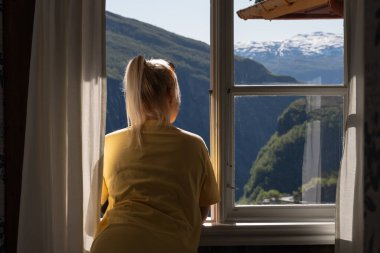 Atkuyruklu sarışın bir kadın pencereden dışarı bakar ve göz kamaştırıcı Norveç fiyortlarına ve karlı dağlara bakar..