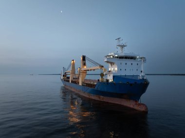 Aydınlatılmış vinçleri olan büyük bir kargo gemisi, alacakaranlıkta, hilal ayının altında, sakin bir denize demirlenir.. 