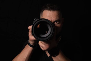 Profesyonel erkek fotoğrafçı, geniş bir kamera lensini izleyiciye izole edilmiş siyah bir arka planda doğrultuyor..
