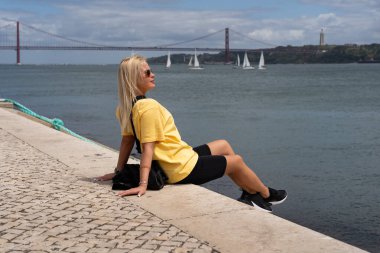 Lizbon 'un rıhtımında dinlenen şık sarışın kadın güneşli günün ve Portekiz' in 25. Abril Köprüsü manzarasının tadını çıkarıyor..