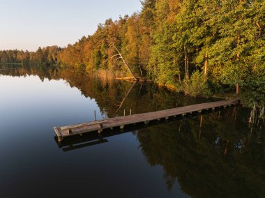 Estonya 'daki sonbahar ormanlarının erken dönemini yansıtan sakin bir gölün üzerindeki sakin bir ahşap iskelenin insansız hava aracı görüntüsü.