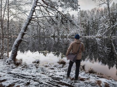 Karlı bir göl kıyısında çantalı bir adam, Estonya 'nın güzel doğasındaki orman yansımasına hayran..