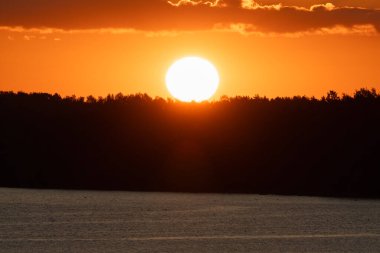 Vibrant Golden Sunset with Large Sun Disk Setting Behind Forest Silhouette Over Calm Lake in Estonia.