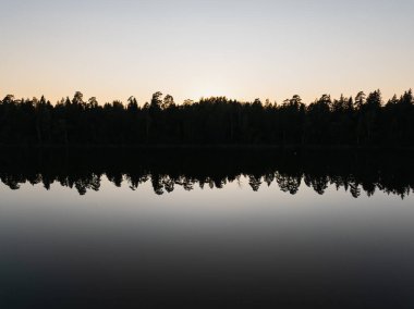 Karanlık orman siluetinin hava aracı görüntüsü Estonya kırsal doğasında sakin gün batımında sakin göl suyuna yansıyor..