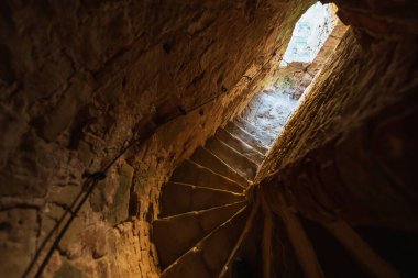Karanlık bir ortaçağ kale kulesinin içine sarılı antik taş sarmal merdiven, yukarıdaki bir açıklıktan aydınlık gün ışığıyla aydınlanıyor, tarihi ve yükselişi vurguluyor..