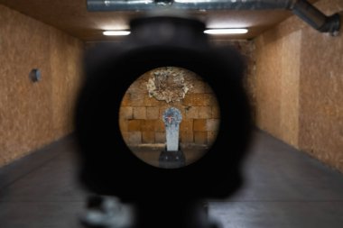 Tüfek dürbününden birinci şahıs görüş mesafesi kırmızı noktalı görüş ile loş ışıklandırılmış, ahşap duvarlı kapalı bir atış poligonunda çelik bir hedefi hedef alıyor..