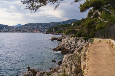 Saint-Jean-Cap-Ferrat 'ın engebeli kayalıklarında Akdeniz ve uzak Fransız Riviera kasabasına bakan sahil yürüyüş yolu..