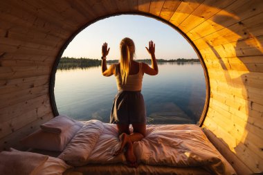 Kadınlar, sakin bir göle bakan yuvarlak pencereli eşsiz bir ahşap kulübeden güzel bir gün doğumunun keyfini çıkarırlar..