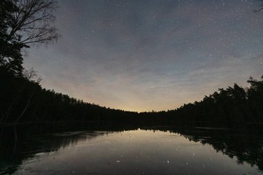 Güzel yıldızlı gece gökyüzü, Estonya doğasında kış boyunca karanlık çam ormanlarıyla çevrili sakin donmuş bir göle yansıyor..