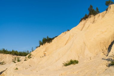 Aydınlık bir günde, açık mavi gökyüzünün altındaki eski bir taş ocağında, dik bir kumlu tepede, disk golf sepeti..