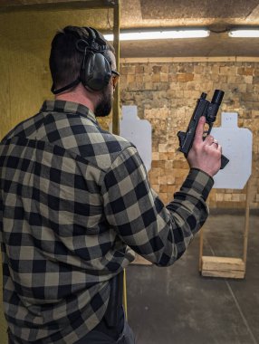 Ekose gömlekli bir adam modern bir tabancayla atış talimi yapıyor. Kapalı alanda kırmızı noktalı bir atış poligonunda..