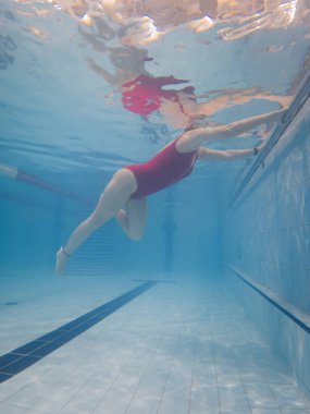 Kırmızı mayolu bir kadının su yüzeyindeki yansımalarıyla havuz kenarına uzandığı dikey sualtı görüntüsü..