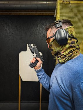 Taktik teçhizat ve yüz maskesi takmış, yarı otomatik modern bir tabancayı kapalı alanda tutan bir adamın yan görüntüsü..
