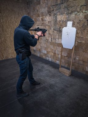 Koyu renk kapüşonlu bir adam, küçük makineli tüfekle atış talimi yapıyor. Kapalı alanda bir hedefe nişan alıyor..