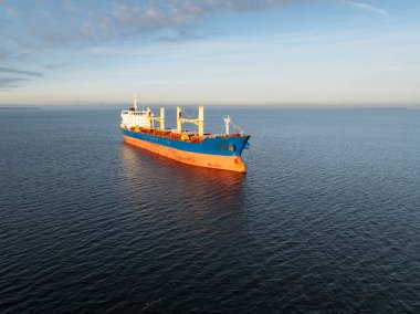 Altın saat boyunca açık denize demir atmış büyük mavi ve turuncu bir kargo gemisinin hava görüntüsü..