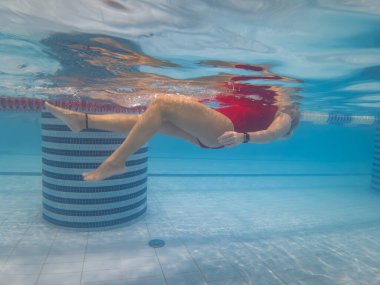 Kırmızı mayo giymiş genç bir kadın açık mavi bir yüzme havuzunda suyun altında yüzerken fayanslı zemine güneş ışığı yansıyor..