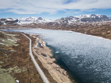 Norveç 'in karlı dağlarında kısmen donmuş bir göl boyunca uzanan dolambaçlı bir yolun hava aracı görüntüsü mavi gökyüzünün altında.