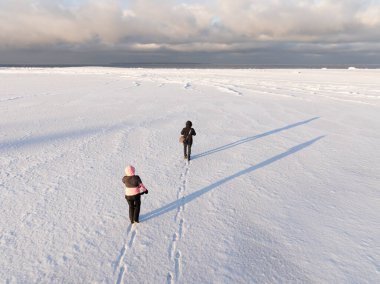 Geniş karlı arazide bulutlu gökyüzünün altında donmuş denize doğru yürüyen iki kişinin yüksek açılı hava aracı görüntüsü..