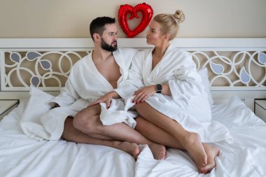 Beyaz bornozlu romantik çift otel odasında yatakta oturmuş, yukarıda kırmızı kalp balonuyla sevgililer gününü kutluyorlar..