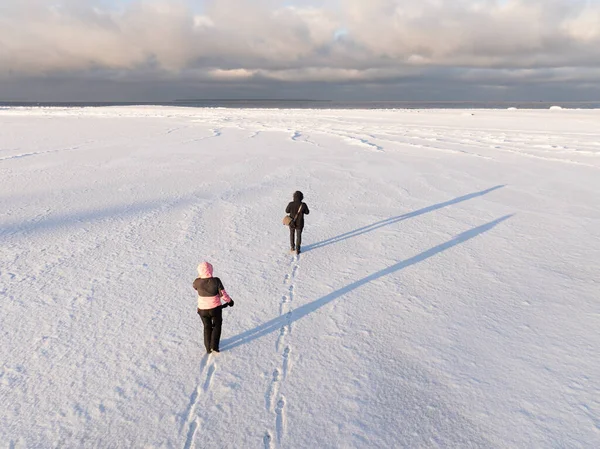 Geniş karlı arazide bulutlu gökyüzünün altında donmuş denize doğru yürüyen iki kişinin yüksek açılı hava aracı görüntüsü..