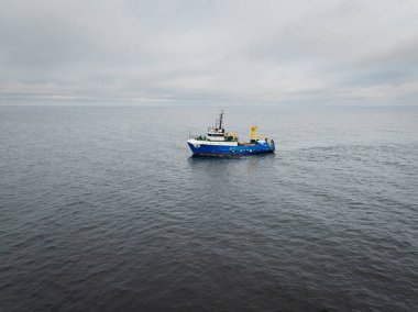 Açık Baltık Denizi 'nde bulutlu bir gökyüzü, endüstriyel balıkçılık endüstrisi ve deniz taşımacılığı altında ağ çeken mavi ticari balıkçı teknesinin insansız hava aracı görüntüsü.