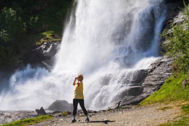 Sarı tişörtlü mutlu sarışın kadın Norveç 'te güçlü bir dağ şelalesinin yanında durmuş manzaralı yaz yolculuğunun ve maceranın tadını çıkarıyor..