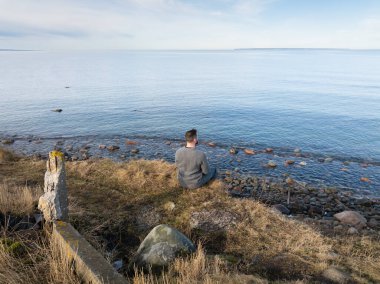 Yüksek açılı drone görüntüsü olan yalnız bir adam çimenli bir uçurumda oturmuş sakin mavi deniz suyuna bakıyor..