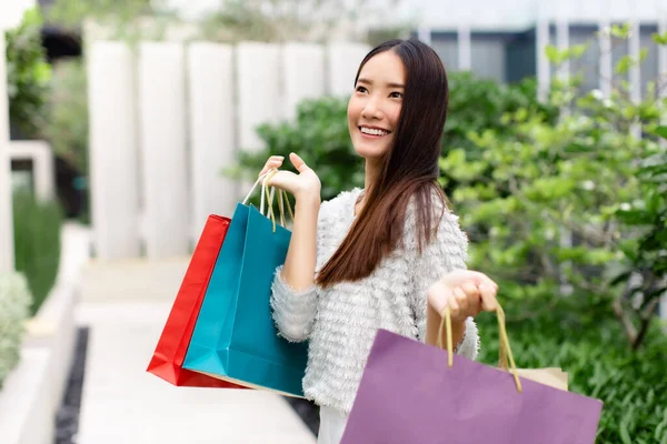 Smiling young Asian woman with shopping colour bags over mall background. using a smart phone shopping online  and smiling while standing mall building. lifestyle shopping in city concept.