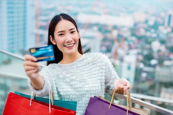 Smiling young Asian woman with shopping colour bags over mall background. using a smart phone shopping online  and smiling while standing mall building. lifestyle shopping in city concept.