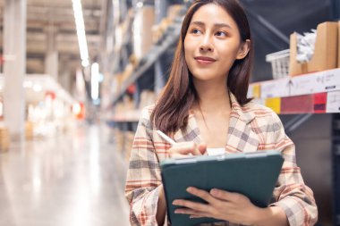 Gülümseyen Asyalı yönetici kadın portresi ayakta duruyor ve depodaki eşyaları kontrol etmek için tablet bilgisayardan ayrıntıları sipariş ediyor..