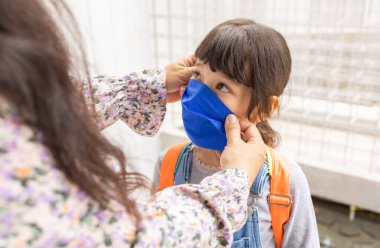 Asyalı anne kızına yardım ediyor ve yüz koruma maskesi takıyor. Küçük kız koronavirüs salgını sırasında okula gidiyor. Anne ve kızı okula gidiyor. Öğrenci çocuk okula gitmeye hazır..