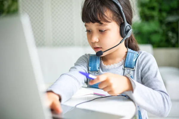 小さな混合レースの女の子は ノートパソコン上の机の研究では スマート小さな子供の手書きノートブック学習では ホームスクーリングの概念にインターネットレッスンを使用して座って ストックフォト