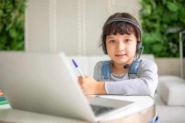 小さな混合レースの女の子は ノートパソコン上の机の研究では スマート小さな子供の手書きノートブック学習では ホームスクーリングの概念にインターネットレッスンを使用して座って ロイヤリティフリーのストック写真