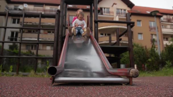 Dos niñas lindas que se divierten mucho mientras cabalgan por el tobogán en el patio — Vídeos de Stock