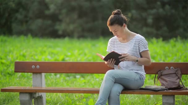 Schöne Frau, die auf einer Bank im Park sitzt und ein Buch liest, Studentin, die sich im Garten auf die Prüfung vorbereitet, junge Frau, die es sich auf einer Parkbank gemütlich macht — Stockvideo