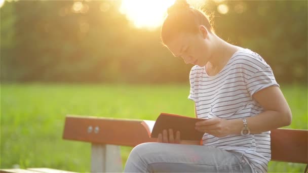 Schöne Frau, die auf einer Bank im Park sitzt und ein Buch liest, Studentin, die sich im Garten auf die Prüfung vorbereitet, junge Frau, die es sich auf einer Parkbank gemütlich macht — Stockvideo