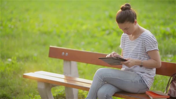 Mulher jovem bonito com tablet no parque, mulher bonita usando um tablet em um banco no jardim, jovem estudante sentada em um banco no parque e usando um tablet — Vídeo de Stock