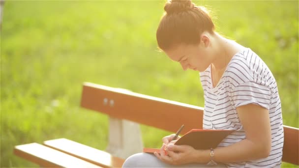 Hermosa joven mujer está escribiendo un diario al aire libre en el parque, estudiante que estudia en un banco en el parque, estudiante chica preparándose para el examen sobre la naturaleza — Vídeos de Stock