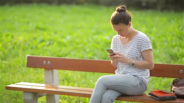 Jonge lachende vrouw met behulp van de smartphone zit op bankje in park. Mooi Europees meisje texting op telefoon — Stockvideo