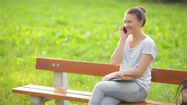 Jovem bela mulher sentada em um banco no parque da cidade e falando ao telefone e sorrindo — Vídeo de Stock