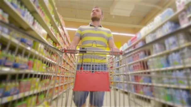 Il ragazzo con il carrello del supermercato pieno si avvicina alla fotocamera e sorride felicemente, il giovane sceglie i prodotti al supermercato, l'uomo compra bevande in negozio — Video Stock