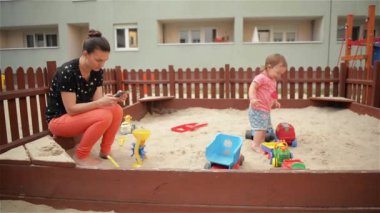 Genç güzel annem kendini kum havuzunda oynayan çocuk kızı, telefon kullanarak anne eğlenirken onun akıllı ve bu iletileri zaman denetler
