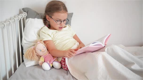 Маленькая девочка в очках листает книгу, пока лежит в постели. Возможно, ребенок болен. Рядом с ней сидит кукла . — стоковое видео