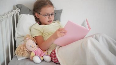 Küçük kız yatakta yatan bir kitap okuma gözlük. Onun yanında bir bebek oturur.