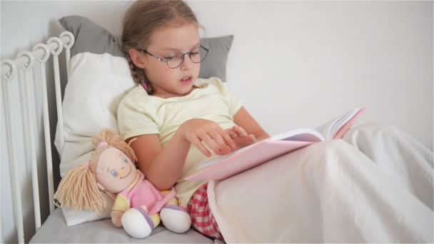 Niña con gafas hojeando un libro mientras está acostada en la cama. Tal vez un niño está enfermo. Junto a ella se sienta una muñeca . — Vídeo de stock