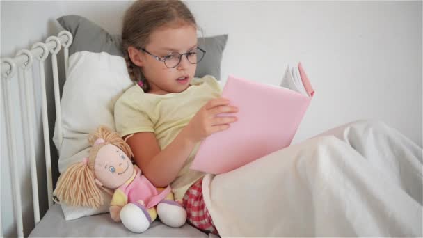 Kislány az üvegek, miközben az ágyban fekve egy könyvet olvas. Mellette ül a baba. — Stock videók